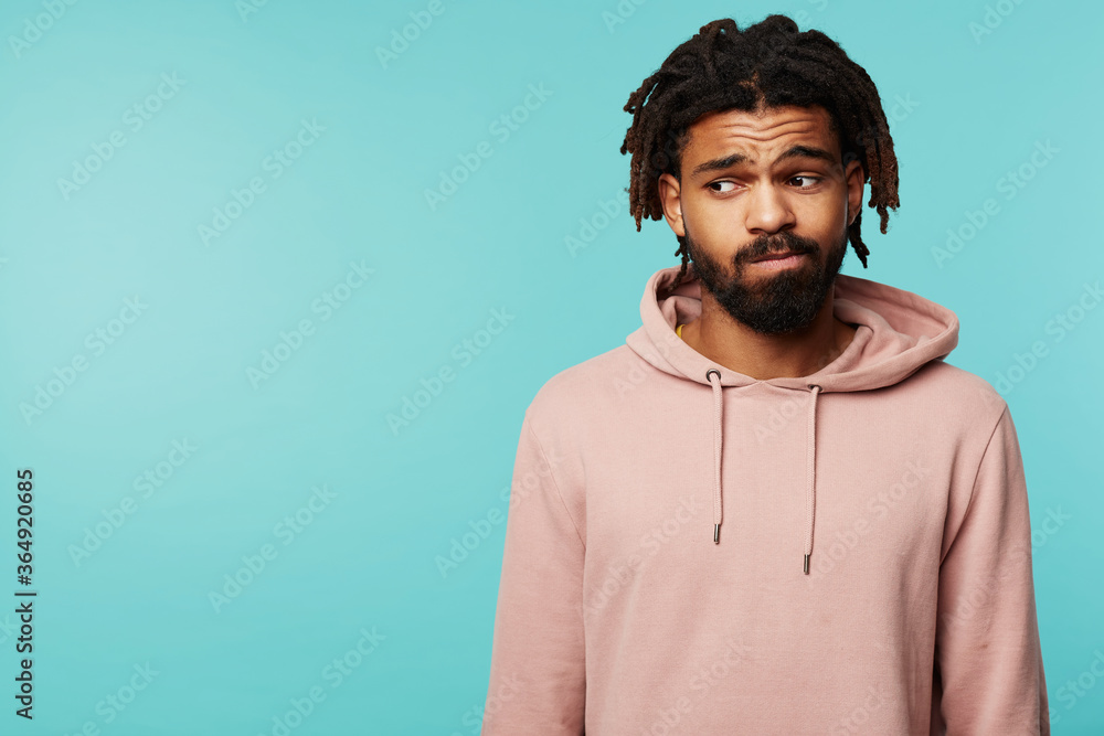 Upset young handsome dark skinned brunette bearded man folding his lips while looking sadly aside, keeping hands down while posing over blue background Upset young handsome dark skinned brunette bearded man folding his lips while looking sadly aside, keeping hands down while posing over blue background