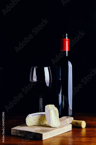 Fototapeta Naklejka Na Ścianę i Meble -  A glass of red wine, a bottle and a piece of cheese on a wooden table and a dark background