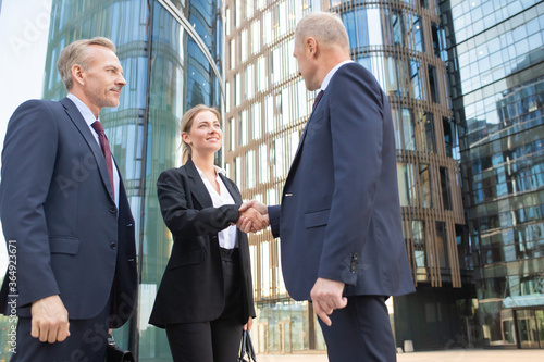 Confident middle-aged businessman greeting young businesswoman and handshaking. Successful content colleagues standing outdoors and glass building on background. Teamwork and partnership concept