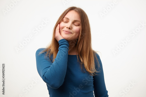 Pleased young attractive blonde lady keeping her eyes closed while smiling gently and touching her cheek with raised palm, standing over white background