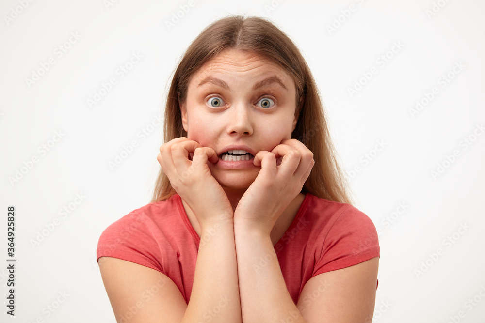 Frightened young green-eyed blonde woman with loose long hair holding her face while looking scaredly at camera, standing over white background in casual clothes Frightened young green-eyed blonde woman with loose long hair holding her face while looking scaredly at camera, standing over white background in casual clothes