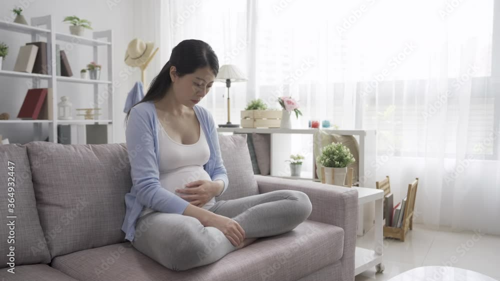 Struggling with morning sickness. Depressed pregnant asian korean woman holding hand cover face and keeping eyes closed while sitting on couch in home living room. future parenthood lady feeling down Struggling with morning sickness. Depressed pregnant asian korean woman holding hand cover face and keeping eyes closed while sitting on couch in home living room. future parenthood lady feeling down