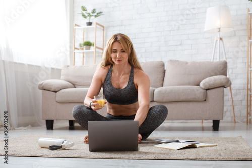 Healthy lifestyle, diet and sport. Woman sitting in lotus pose on floor with glass of juice, looking at laptop