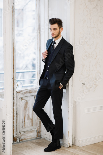 groom in a black suit in a photo studio. posing near the window. elegant wedding © Denys