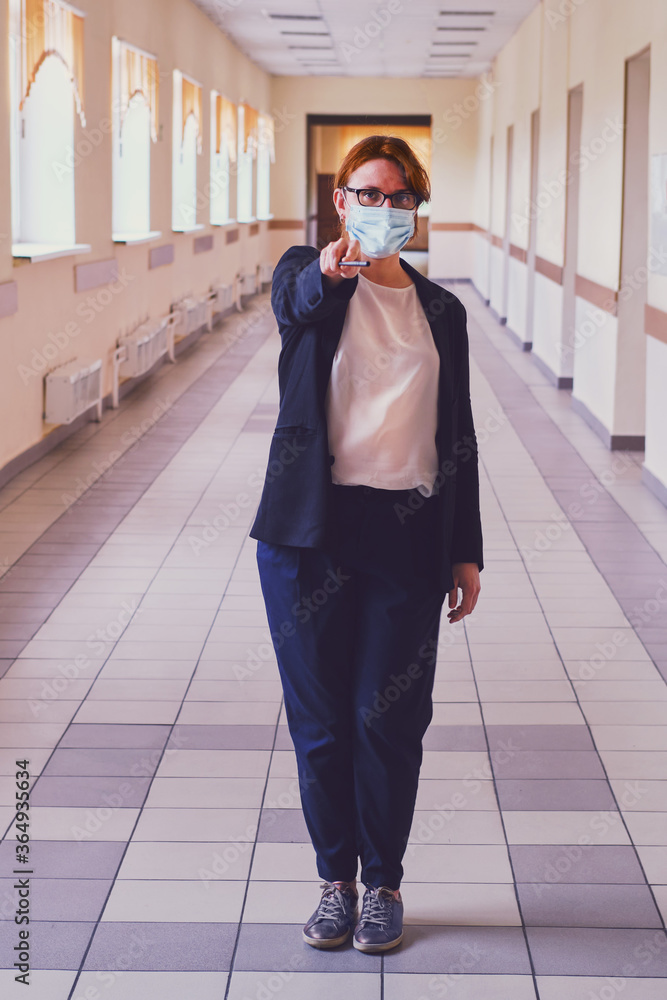 Fototapeta premium Female teacher in face mask stands in an empty school corridor. Coronavirus quarantine concept