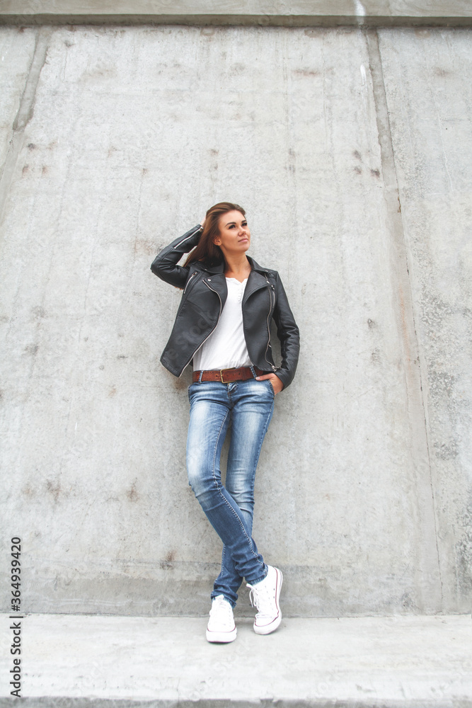 Obraz premium Young cute woman is standing by the wall in a leather jacket, jeans and sneakers.