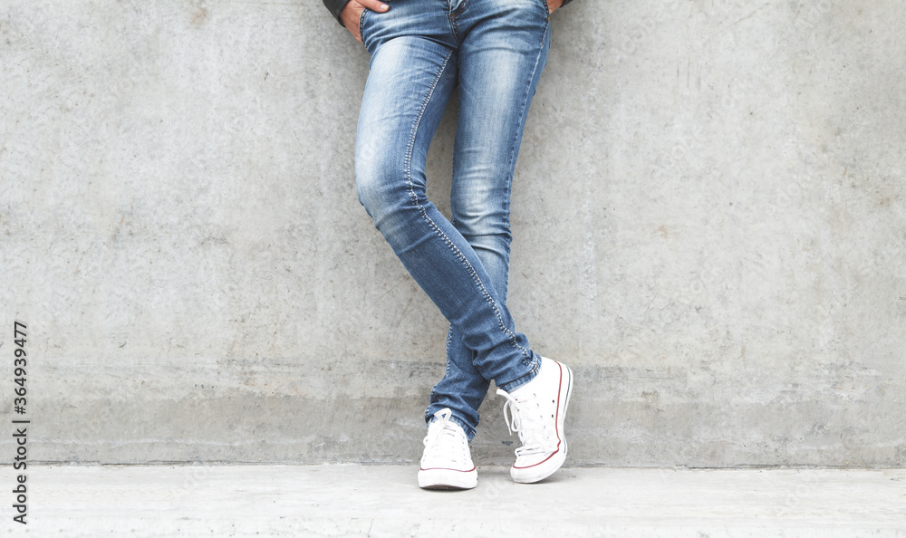 Shapely female legs in sneakers and jeans near a concrete wall.
