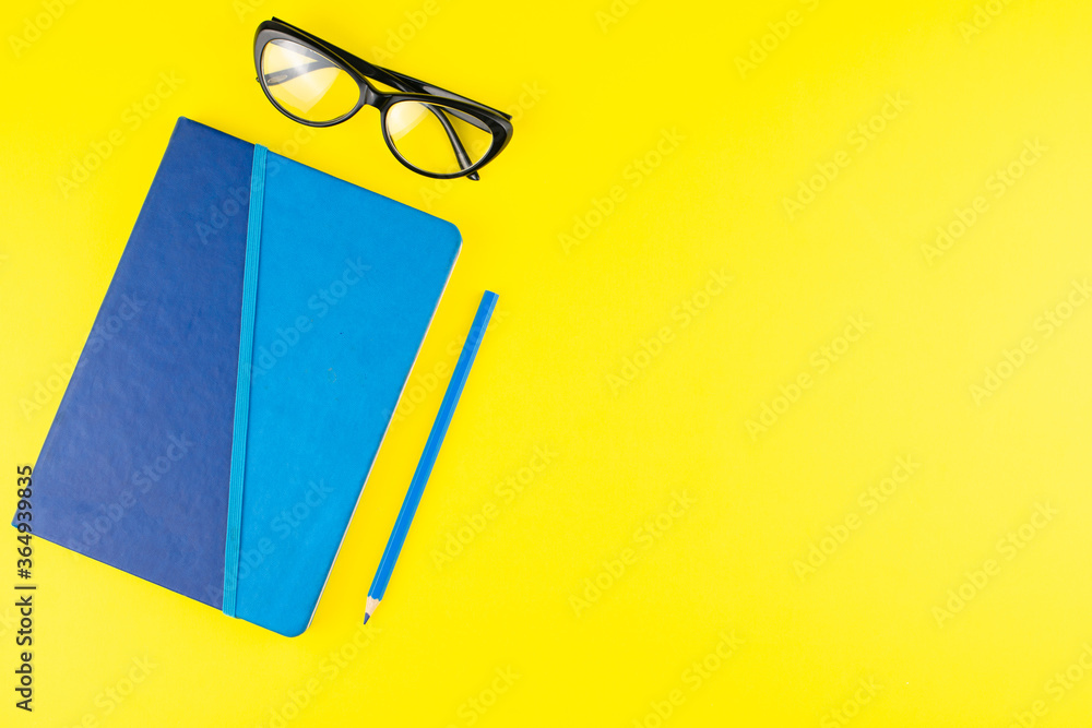 Blue notepad and pencil, black glasses on yellow background. Flat lay. Copy space. Workplace in the office. Blue notepad and pencil, black glasses on yellow background. Flat lay. Copy space. Workplace in the office.