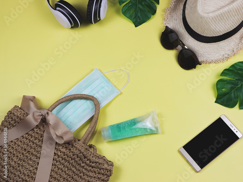 COVID-19 prevention , travel and new normal concept, top view of woven bag with surgical mask and alocohol gel and women's vacation accessories on yellow background.