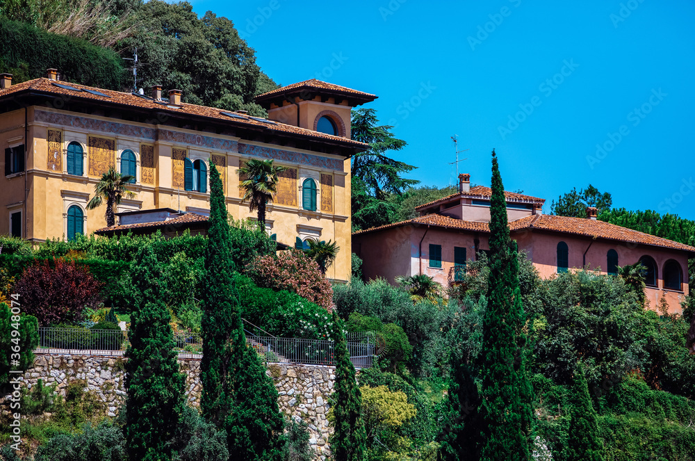 Beautiful and old houses on the hillside, surrounded by trees and green bushes. Blue and clear sky above the houses. stone wall in front of souses