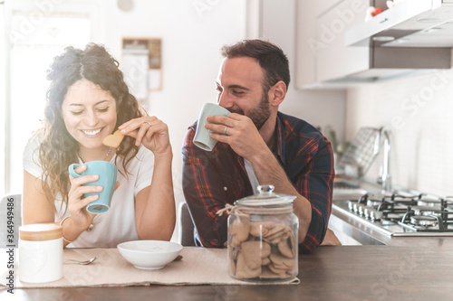 Engaged couple has breakfast together in their new home - young couple smiling while drinking and eating in the kitchen - warm filter on background