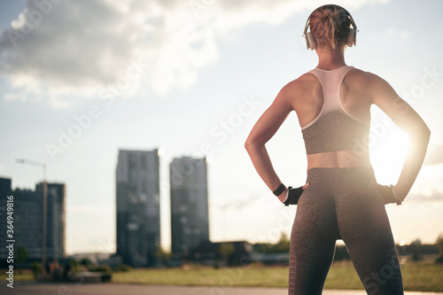 Back view of strong fitness woman in sportswear and headphones, back light, city view