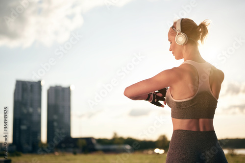 Fototapeta Naklejka Na Ścianę i Meble -  Back view of strong muscular woman in sportswear and headphones is cheking smartwatch and using mobile application, back lit, city view