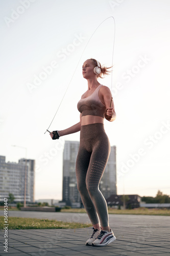 Strong muscular sporty woman in headphones doing physical exercise using jump/skipping rope in summer park. Fitness workout. Sport, healthy lifestyle.