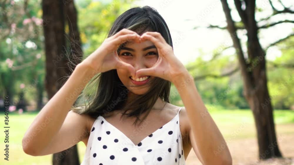 Young brunette woman with smile face making heart shape hands gesture in park. Beautiful Asian girl with toothy smile outdoors. Young brunette woman with smile face making heart shape hands gesture in park. Beautiful Asian girl with toothy smile outdoors.