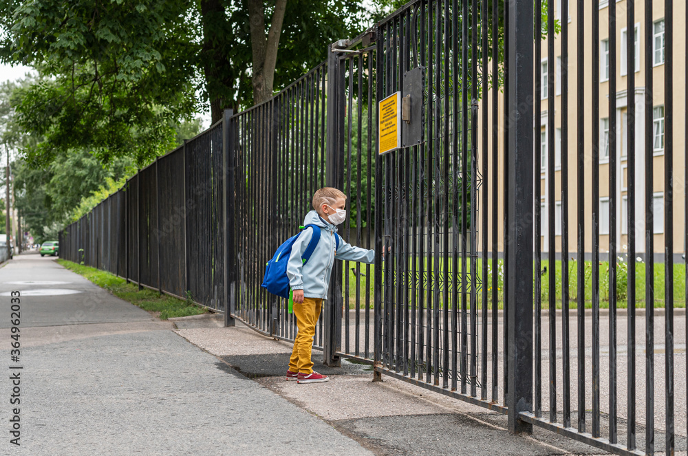 A masking kid is going to school for the first time. A boy with a bag goes to primary school. A student with a backpack open gate to school. Back to school.