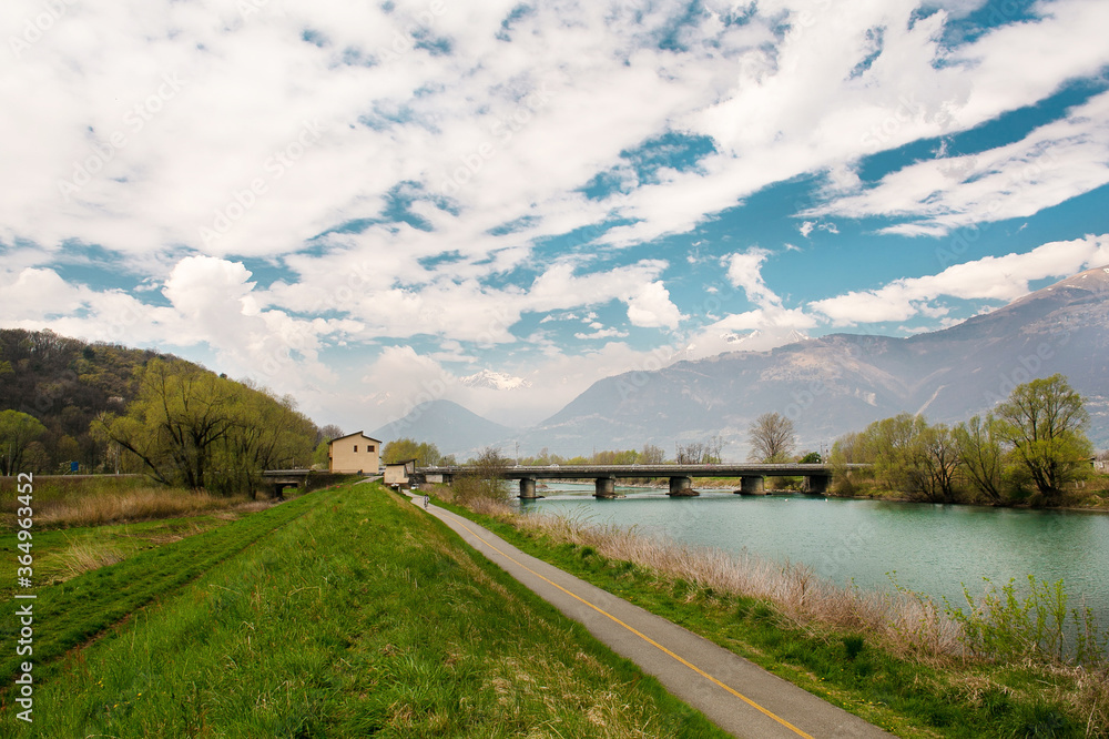 Railway bridge over the Ada river in Lombardy. Blue water, trees and mountains on the background. Clouds, shadows, rockes and stones in water. Distance view. Green grass and road 
in the foreground.