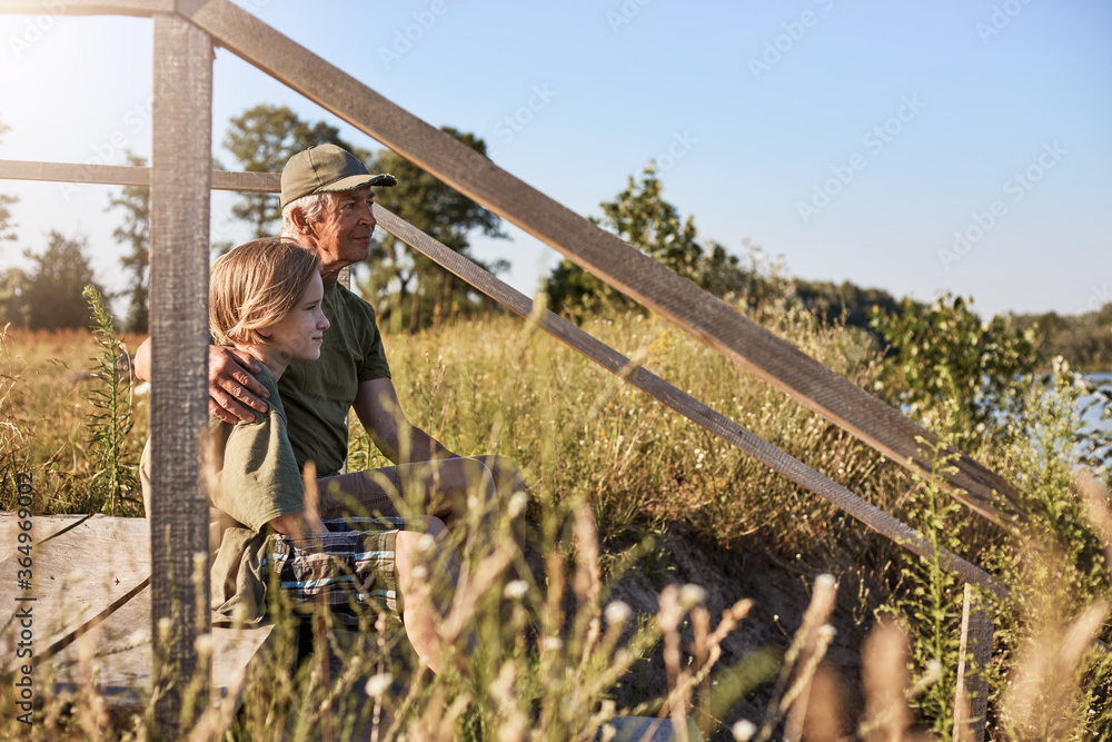 Elderly man sitting on wooden placing with his son or grandson and hugging him, looking far away, enjoying beautiful nature, posing in meadow, spending time together. Elderly man sitting on wooden placing with his son or grandson and hugging him, looking far away, enjoying beautiful nature, posing in meadow, spending time together.