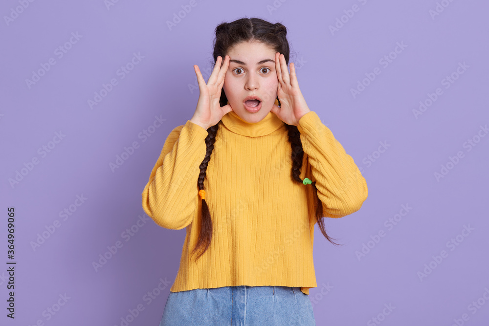 Stunned dark haired woman with two pigtails, keeping hands near face, afraids of something astonishing, dresses in fashionable yellow shirt, posing isolated over lilac background. Stunned dark haired woman with two pigtails, keeping hands near face, afraids of something astonishing, dresses in fashionable yellow shirt, posing isolated over lilac background.