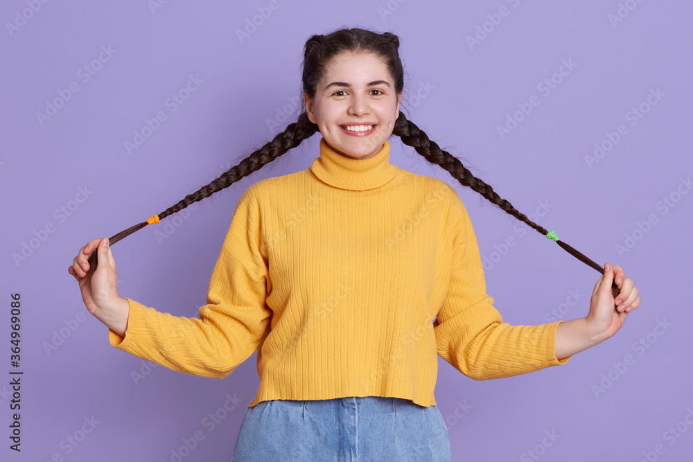 Happy charming lady with two pigtails looking directly at camera and playing with her hair, posing isolated over lilac background, girl with broadly smile.