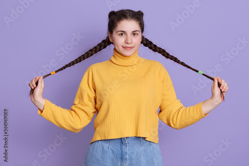 Good looking brunette woman holds two pigtails in hands, looks at camera with satisfied expression, wears stylish yellow jumper, girl smiles pleasantly,