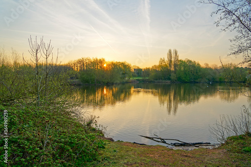 Spring sun rises behind tall trees along the banks of a small lake