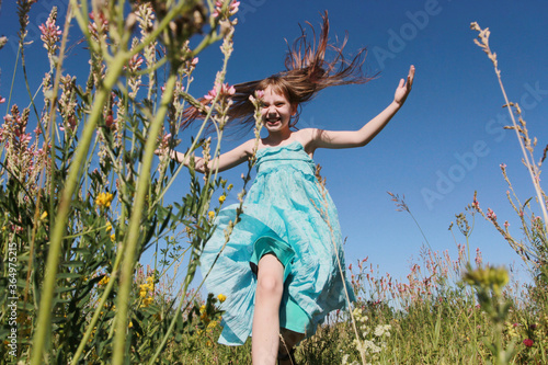 Cheerful happy girl running through flowery meadow or field, having fun, making flying hair, spreading arms. Low angle. Country leisure time, summer vacation, euphoria, lifestyle, childhood concept