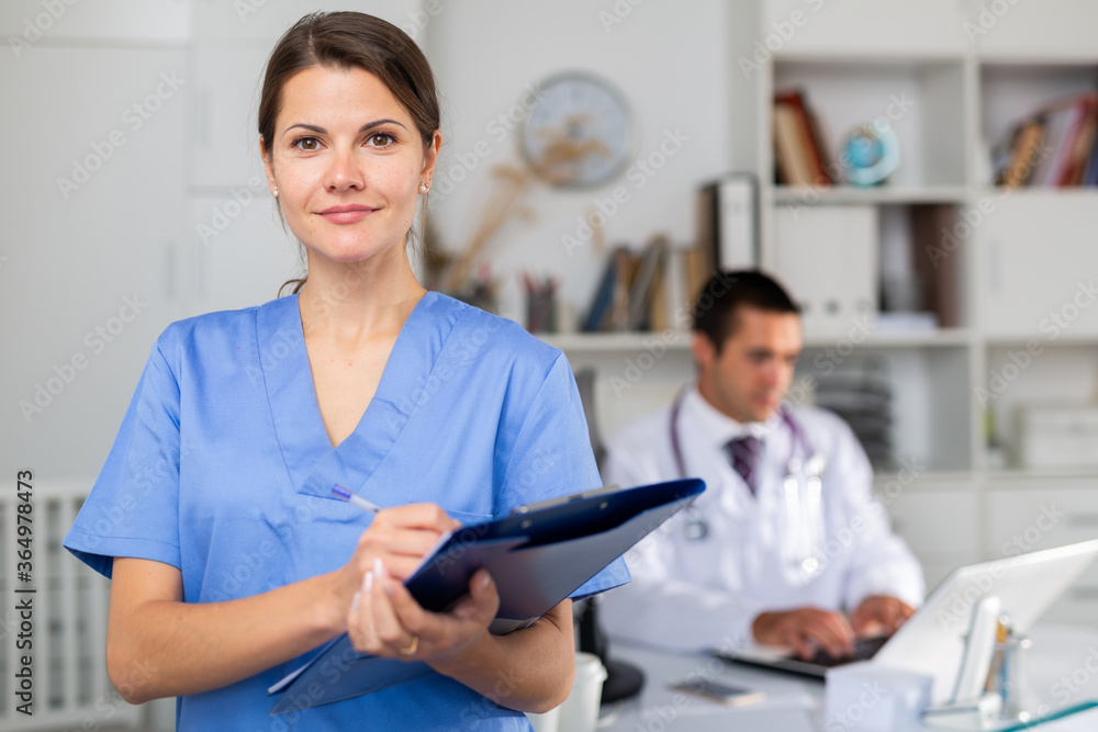 Positive brunette woman doctor standing in office with clipboard in hands, writing medical history sheet..