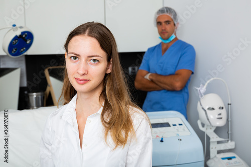 Portrait of young woman client of beautician sitting in clinic waiting for procedures, man doctor on background