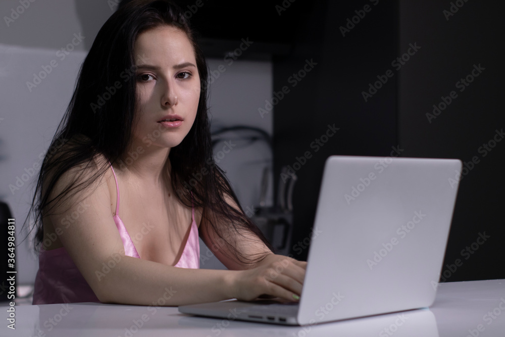 tired, pensive, beautiful brunette girl in pink pajamas with a blue tea-coffee cup, works on a laptop - remote work or study - in a gray kitchen. High quality photo tired, pensive, beautiful brunette girl in pink pajamas with a blue tea-coffee cup, works on a laptop - remote work or study - in a gray kitchen. High quality photo