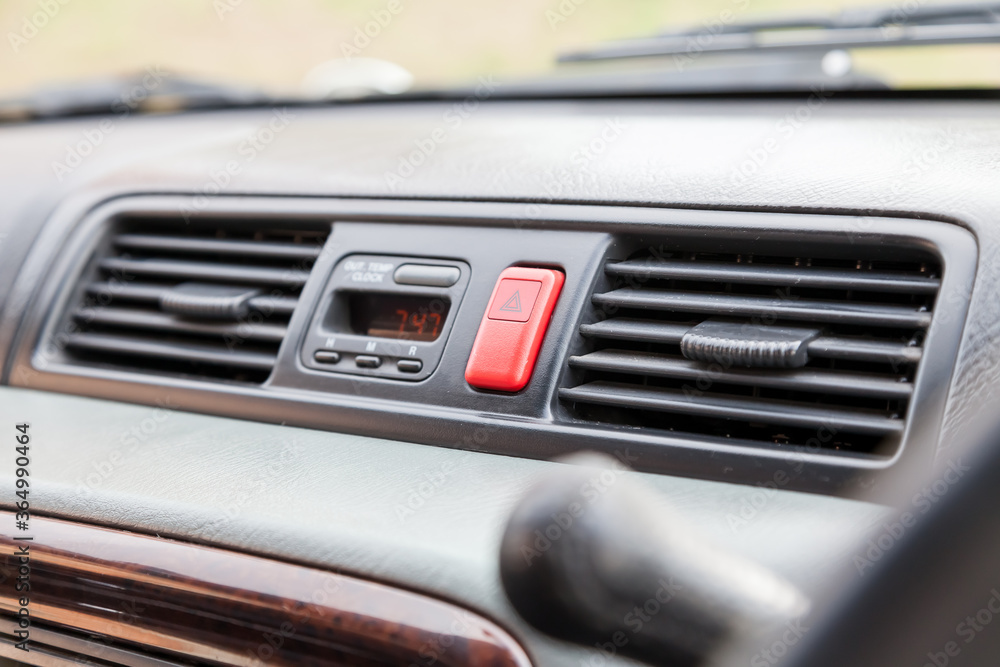 Closeup of a red emergency light button with a white triangle between the air conditioning grilles on the dashboard in the interior of a car.