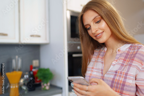 Pretty young woman in casual clothes using her smartphone in kitchen