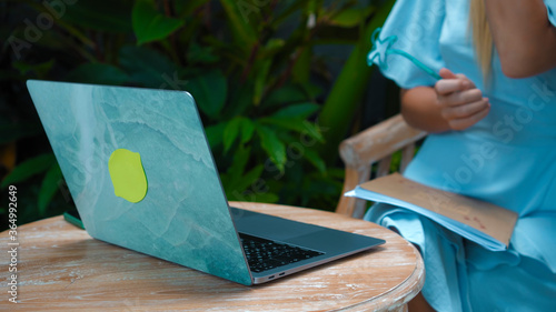 A girl in a blue dress remotely online working behind a laptop showing her thumbs in the backyard with green sprouts on the background
