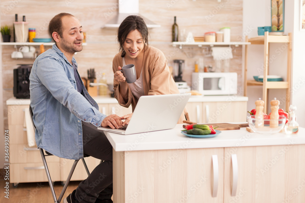 Entrepreneur working on laptop in kitchen and wife looking at his work. Happy loving cheerful romantic in love couple at home using modern wifi wireless internet technology Entrepreneur working on laptop in kitchen and wife looking at his work. Happy loving cheerful romantic in love couple at home using modern wifi wireless internet technology