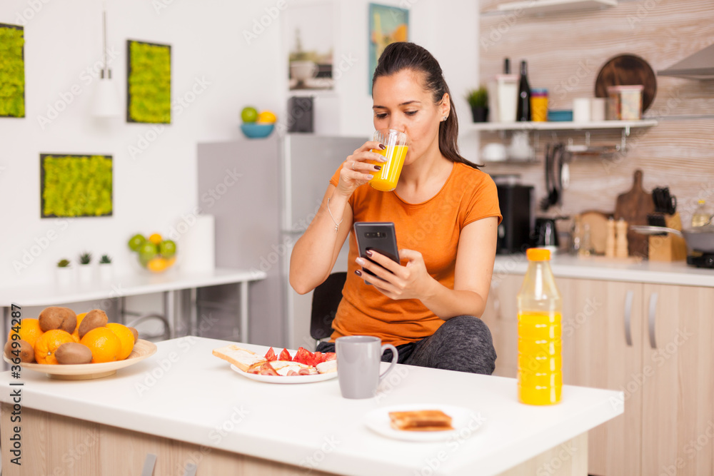 Young freelacer looking in the phone while drinking ogange juice. Housewife using modern tehcnology and drinking healthy, natural, homemade orange juice. Refreshing morning Young freelacer looking in the phone while drinking ogange juice. Housewife using modern tehcnology and drinking healthy, natural, homemade orange juice. Refreshing morning
