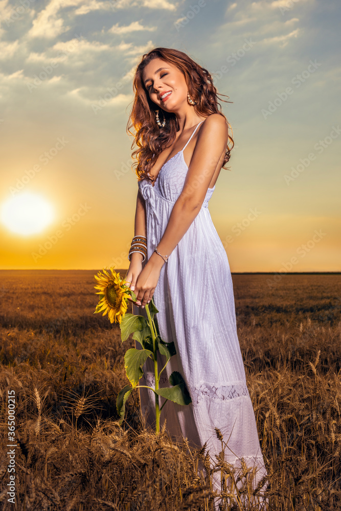 Obraz premium Beautiful woman in white dress in a wheat field with a sunflower flower at sunset.