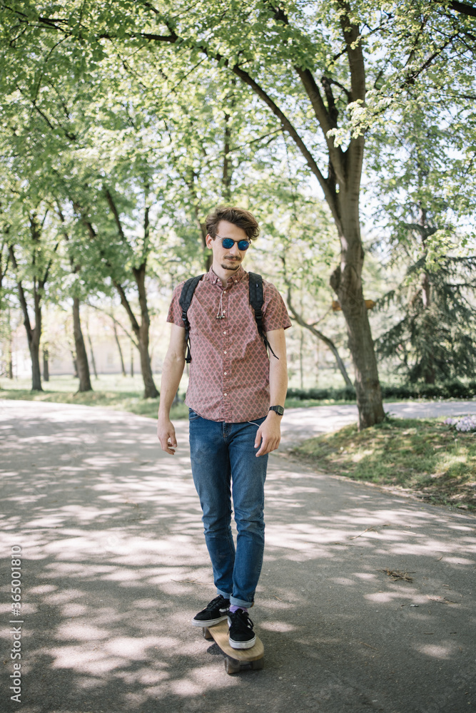 Guy skateboarding on park road. Cool man dressed in casual outfit riding skateboard on shadowed road in park during sunny day. Guy skateboarding on park road. Cool man dressed in casual outfit riding skateboard on shadowed road in park during sunny day.