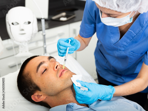 Young man patient of beautician receiving rejuvenating facial injections caring of his skin
