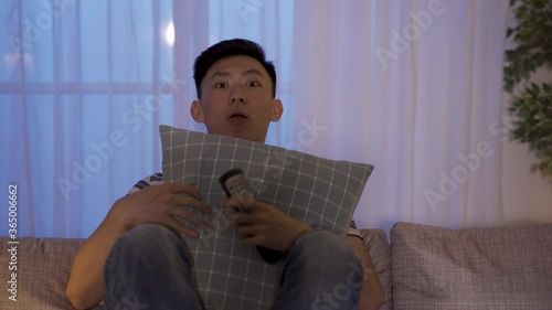 chinese male huddling up on couch with pillow is seeing thriller at wee hours. asian man staring at screen is scared by scary ghost and yelled, but continues watching after catching his breath.