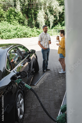 Selective focus of smiling man holding coffee to go near wife while fueling auto on gas station
