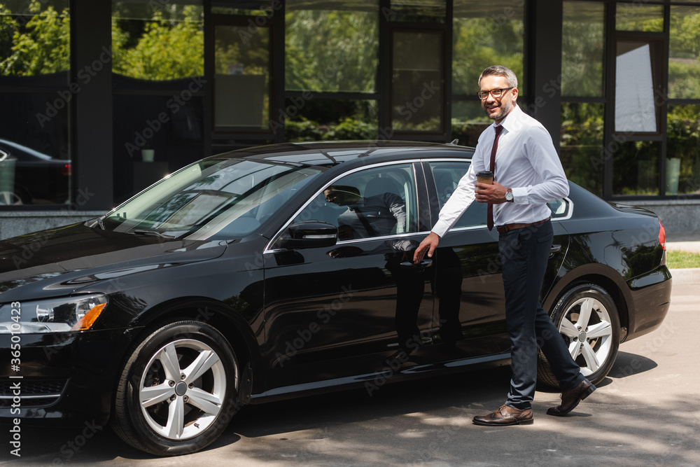 Smiling businessman holding coffee to go near car on urban street Smiling businessman holding coffee to go near car on urban street