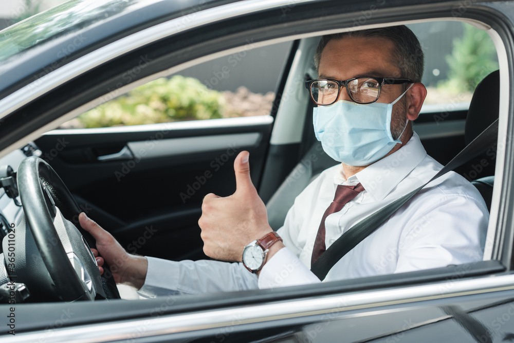 Selective focus of businessman in medical mask showing thumb up while driving auto Selective focus of businessman in medical mask showing thumb up while driving auto