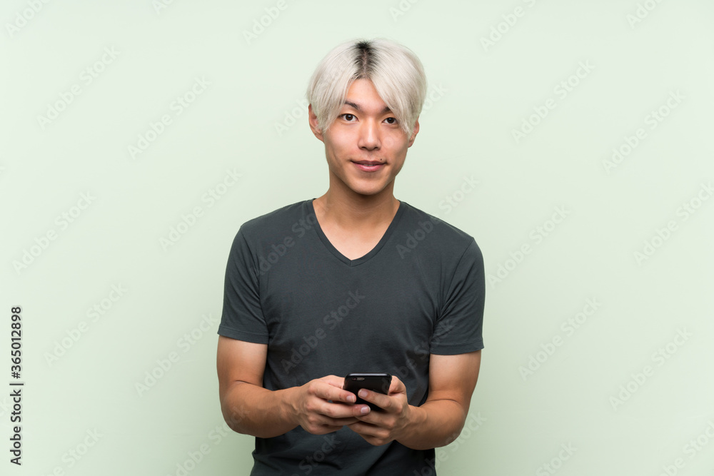 Young asian man over isolated green background sending a message with the mobile Young asian man over isolated green background sending a message with the mobile