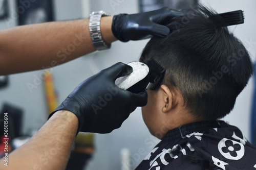 hairdresser cutting hair of a client. An Asian boy is sitting in a barber shop.