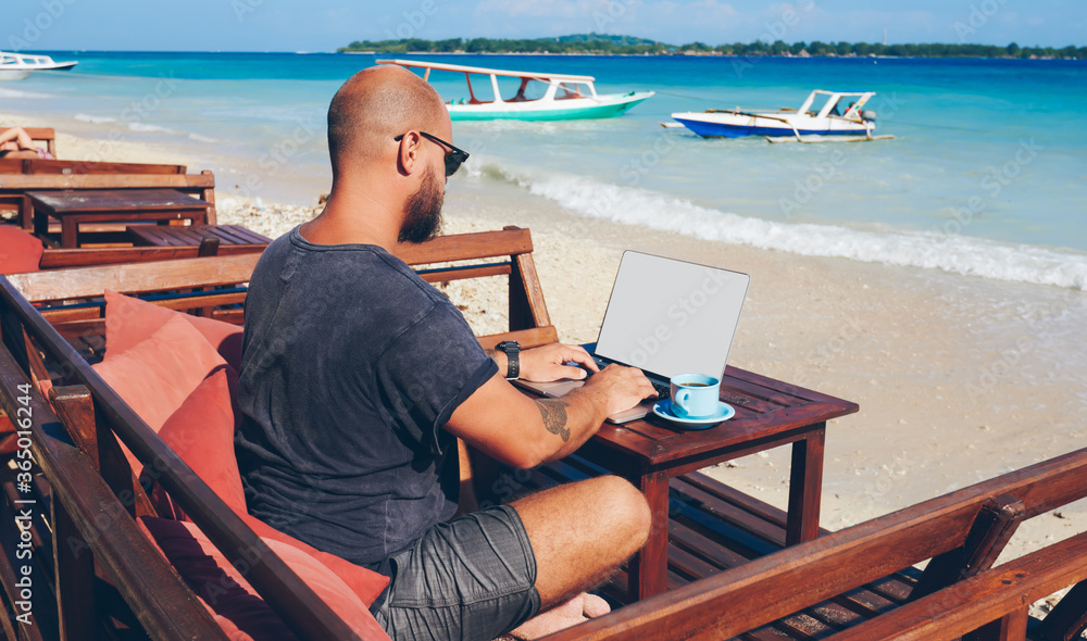 Skilled male freelancer working online on his business via laptop computer while resting on ocean shore, millennial hipster guy update content in his blogg in keyboarding publication about trip