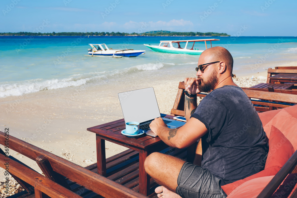 Pensive male freelancer thinking about idea for new article recreating on ocean shore with laptop computer,businessman using netbook with mock up screen for remote job during vacation on resort.