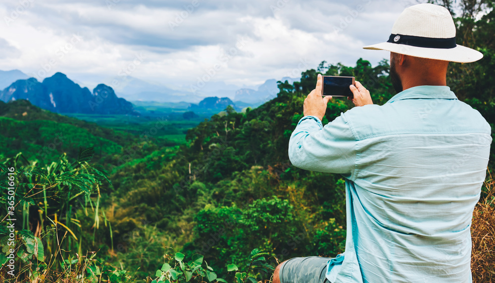Back view of a man wanderer is shooting video on smart phone, while is sitting on a mountain against subtropical forest. Young hipster guy is taking photo on cell telephone of a beautiful jungle view