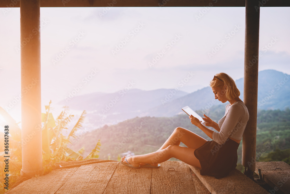 Female traveler is watching video on digital tablet,while is sitting against amazing subtropical forest and sky background with copy space. Woman tourist is searching information on portable touch pad