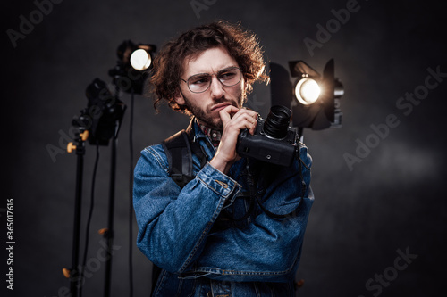 Young stylish man photographer with curly hairstyle in glasses holds digital camera with hand on chin, looks thoughtful. Dark photo studio with lighting equipment in the background