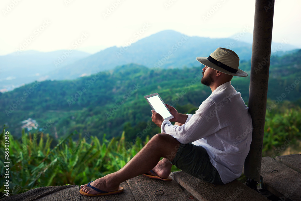 Dreaming man is holding touch pad with copy space on screen, while is enjoying amazing jungle view. Male author is admiring beautiful Asian scenery, while inspire to write his book via digital tablet